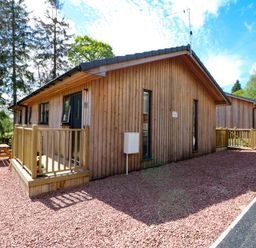 Wooden cabin with fenced porch and picnic table on gravel surrounded by trees at Broken-Sky Lodge in Otterburn