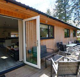 An outdoor wooden deck with seating and a table next to a wooden building with open glass doors at Broken-Sky Lodge in Otterburn