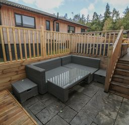 An outdoor seating area with a grey wicker corner sofa and table surrounded by wooden fencing and decking at Barn Owl in Otterburn