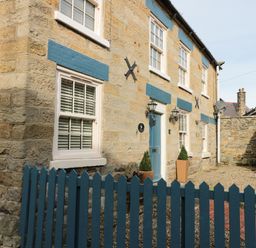A stone house with blue window shutters a blue door fenced front yard and two potted plants at The Nook at 29 High Street Cloughton near Burniston