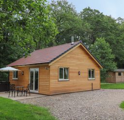 A wooden cabin with a patio table umbrella and chairs near trees and a shed in the background at Park Brook Dell in Scorton