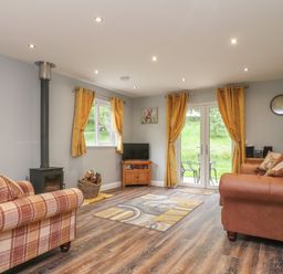 A living room with a wood stove armchairs a TV on a wooden stand and yellow curtains at Park Brook Dell in Scorton
