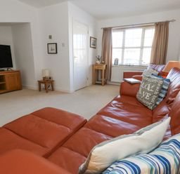 A living room with an orange leather sofa pillows a wooden TV stand with a television a small side table lamp and window with curtains at Fairways in Wyke Regis