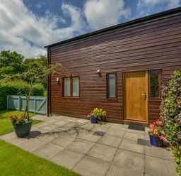 An exterior wooden wall with a wooden door and two windows a paved patio with potted flowers and a small tree a green lawn and a light green fence with trees in the background at Cholwell Barn Apartment Mary Tavy