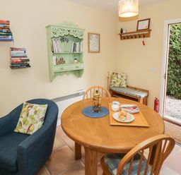 A dining area with a round wooden table set with food and a French press a blue armchair with a cushion a wooden bench with a cushion bookshelves on the wall and a door leading outside at The Garden Lodge in Upton Cressett near Bridgnorth