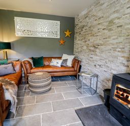 A living room with two leather sofas a round patterned ottoman a side table and a wood burning stove at Swn yr Afon in Beddgelert