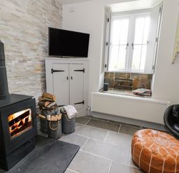 A room with a wood burning stove next to a television on a cabinet and a window seat with books at Swn yr Afon in Beddgelert