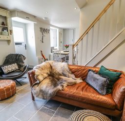 A living room with a brown leather sofa with pillows and a fur throw a black chair with a blanket and cushion a brown ottoman and a dining table with chairs near a staircase at Swn yr Afon in Beddgelert