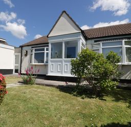 A single story house with a bay window and porch surrounded by a grassy garden with bushes and flowers at Garden View in Quintrell Downs near Newquay