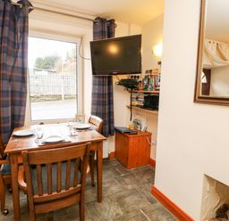 A dining area with a wooden table set for three near a window with blue checked curtains and a wall-mounted TV at Hunter Cottage in Haltwhistle near Hadrians Wall