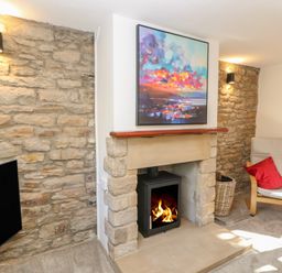 A room with a stone fireplace with a fire burning a painting above it a wooden chair with a red cushion and a basket at The Forge in Allendale