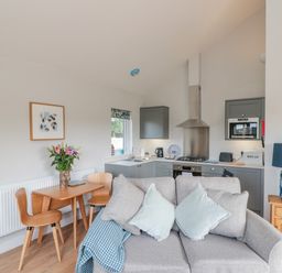 A compact kitchen and dining area with a small wooden table and chairs a grey sofa with cushions and a wooden side table with a blue lamp at Peep o Day in Kirkbymoorside
