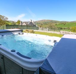 An outdoor hot tub with bubbling water and part of its cover open in a garden with a picnic table and green hills in the background at Bwthyn Y Bugail in Pedairffordd near Llanrhaeadr-Ym-Mochnant