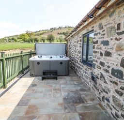 An outdoor patio area with a hot tub next to a stone wall and green railing with countryside view at Y Granar Llanrhaeadr-Ym-Mochnant
