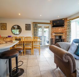 An open living and dining area with a brown sofa and blue cushions a dining table with wooden chairs a brick fireplace with a mounted TV and large windows at Y Stabl (The Stable) in Llanrhaeadr-Ym-Mochnant