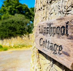A wooden sign reading Washpool Cottage on a stone wall with greenery and a road in the background at Washpool in Milton-Under-Wychwood
