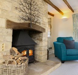 A stone fireplace with a wood-burning stove a basket of firewood a blue armchair and floral curtains at Washpool Milton-Under-Wychwood