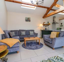 A living room with grey sofas, a wooden coffee table on a round rug, and a kitchen area with exposed wooden beams at Plough Cottage in Little Kelk near Bridlington