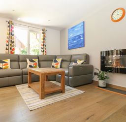 A living room with a grey sectional sofa and patterned cushions a wooden coffee table striped rug a wall clock a potted plant a blue painting and a wall-mounted fireplace at 32 Trevithick in Hayle
