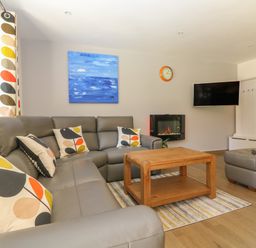 A living room with a gray sectional sofa with patterned cushions a wooden coffee table a wall-mounted television and a fireplace at 32 Trevithick in Hayle