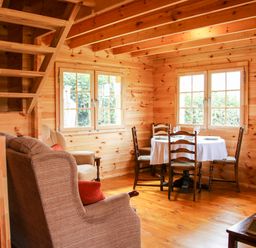 A wooden dining area with a round table covered by a white tablecloth and four chairs next to two windows in a log cabin