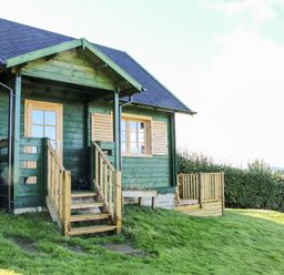 A green wooden cabin with wooden door and windows shutters on a grassy area at Callow Fold in Dinchope near Craven Arms