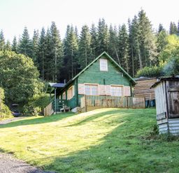 A green wooden cabin with a fenced porch on a grassy slope surrounded by trees and a gravel driveway with a parked car at Callow Fold in Dinchope near Craven Arms