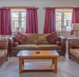 A living room with three brown sofas with plaid cushions red curtains on two windows and a wooden coffee table at Grubbings Graythwaite near Hawkshead