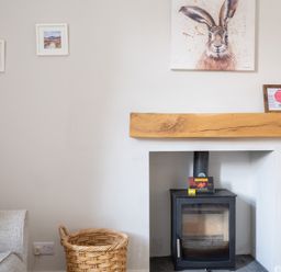 A fireplace with a black stove and a wooden mantle with a rabbit painting above and a wicker basket next to a gray sofa at Briar in Graythwaite near Hawkshead