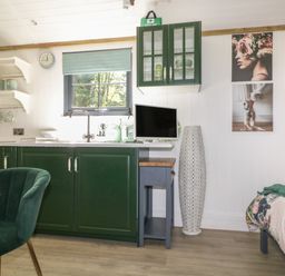 A kitchen area with green cabinets a window with a shade a sink and appliances next to a dining table and a bed at High Rigg Shepherd's Hideaway in Brampton Cumbria