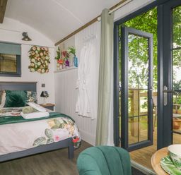 A bedroom with a bed floral bedding robes on hooks and glass doors opening to a deck with chairs at High Rigg Shepherd's Hideaway in Brampton Cumbria