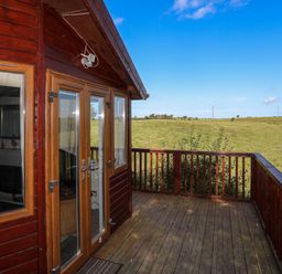 A wooden deck with railing outside a cabin overlooking grassy fields at Ffrwd Lodge in Rhosneigr