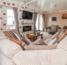 A living room with a large sectional sofa and coffee table a wall-mounted TV and fireplace with floral wallpaper at Jonstone Pines in Cayton Bay near Scarborough