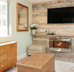 A living room with a wooden TV wall a mounted flat screen TV a wooden cabinet a wooden coffee table a beige ottoman a mirror and a window at Bluebell Wood in Brandesburton