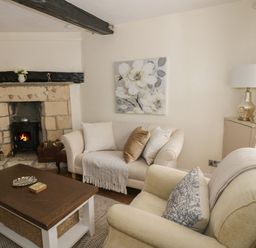 A living room with a stone fireplace two sofas a wooden coffee table and a painting of flowers at Thimble Cottage in Winchcombe