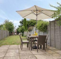 A patio area with a table and four chairs under a large umbrella in a garden with grass and fences on both sides at Thimble Cottage in Winchcombe
