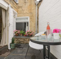 A small outdoor patio with a glass table set with a drink and flowers next to white walls and an open door with potted plants by the window at Thimble Cottage in Winchcombe