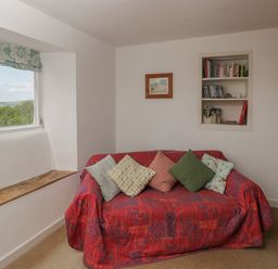 A small sitting area with a red covered sofa and cushions near a window with a wooden sill and built in bookshelf at Athelstan Cottage in Chloe Mead near Stroud