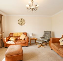 A living room with two brown leather sofas a matching ottoman a green recliner chair a wooden side table with a lamp a wall clock curtains and a framed picture at Glanaig Morfa Bychan