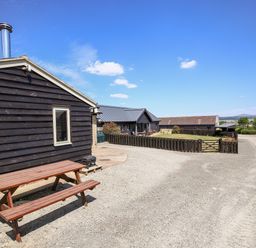 An outside area with a wooden picnic table beside a black wooden building and a gravel driveway at Norton Cottage in Craven Arms