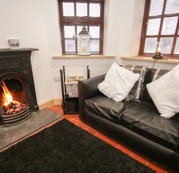 A room with a black leather sofa with white pillows a fireplace with a fire and windows with wooden blinds at Old Tollgate in Gwalchmai near Llangefni