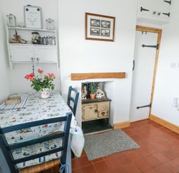 A small dining area with a table covered by a patterned tablecloth and flowers in a vase a shelf with kitchen items a white door and a small stove in a fireplace at Old Tollgate Gwalchmai near Llangefni