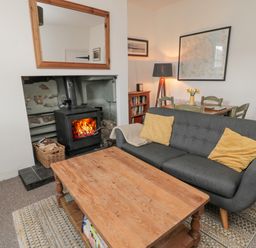 A living room with a wood stove, a grey sofa with yellow cushions, a wooden coffee table, a dining table with chairs, a floor lamp, and framed pictures on the walls at Lavender Cottage in Glanton