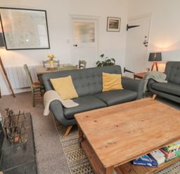 A living room with gray sofas yellow cushions a wooden coffee table board games under the table a fireplace a dining table with green chairs and a floor lamp at Lavender Cottage in Glanton