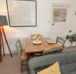 A dining area with a wooden table and four chairs a floor lamp a framed map on the wall a plant on a stand and a gray sofa in Lavender Cottage in Glanton
