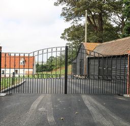 A black metal gate with brick pillars at the entrance of a driveway leading to a white house and black outbuildings at Church Farm in Burgh Castle