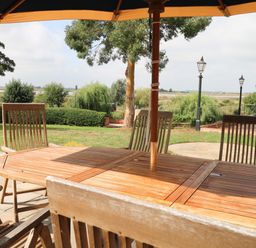 A wooden outdoor dining table with chairs under an umbrella in a garden with trees and lamp posts at Waveney View Cottage in Burgh Castle