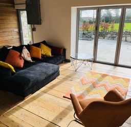 A living room with a black sofa with multi-colored cushions a brown armchair a glass coffee table and a patterned rug in front of large sliding glass doors at The Corn Store in St Stephen