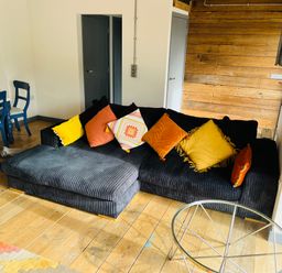 A living room with a black sectional sofa with colorful cushions a glass round coffee table wooden floor and a dining table with blue chairs at The Corn Store in St Stephen