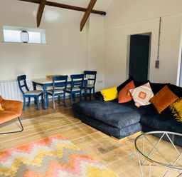 A living room with a dark sectional sofa with colorful cushions a blue dining table with chairs an orange armchair and a patterned rug on wooden floors at The Corn Store in St Stephen